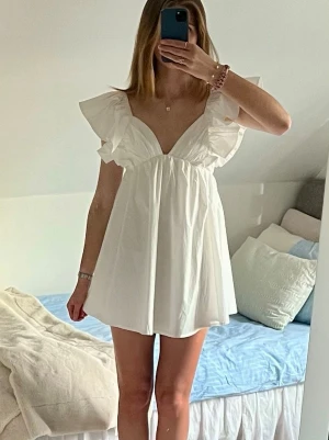 Vit miniklänning med volangärmar - Supersöt vit kort klänning med djup v-ringning, volangärmar och rosett i ryggen💕 Klänningen har en luftig passform och är perfekt för varma dagar☀️ Fungerar också jättebra som studentklänning! Klänningen är helt ny med prislapp, den är endast provad.