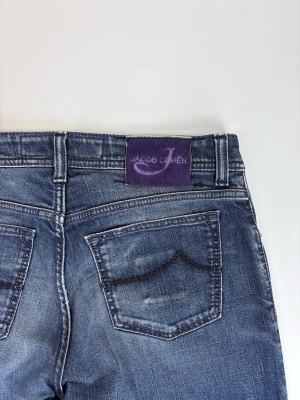 Blå jeans från Jacob Cohen - Snygga blå jeans från Jacob Cohen med klassisk femficksmodell och raka ben. Jeansen har en tvättad look med slitningar och en lila patch med logga bak i midjan. Tillverkade i mjuk denim som ger en skön passform och stilren vibe.
