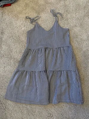 Randig blåvit sommarklänning - En kort klänning med smala blå och vita ränder, knytband på axlarna och volangliknande lager. Klänningen har en avslappnad passform.