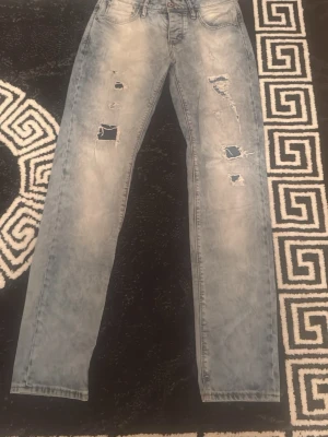 Ljusblå ripped jeans från Genistar - Säljer ett par ljusblå jeans från Genistar med slitna detaljer och flera hål på framsidan. Jeansen har rak passform och klassisk femficksdesign. Perfekta för dig som gillar en avslappnad och trendig stil.