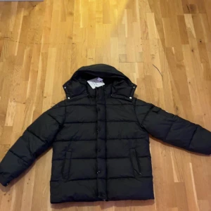 Svart pufferjacka från Moncler verzere - Svart pufferjacka från Moncler med huva och raka, breda kanaler. Jackan har långa ärmar, två sidofickor med dragkedja och knappar framtill. Insidan har klassisk Moncler-etikett och serietidningsmotiv. Perfekt för kalla dagar. Storlek M men passar även L.