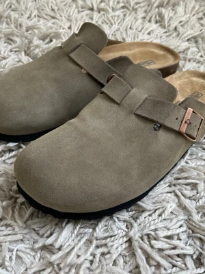 Birkenstock Boston tofflor mocka - Säljer ett par klassiska Birkenstock Boston tofflor i beige mocka med justerbart spänne i metall. De har en mjuk innersula och svart yttersula, perfekta för chill dagar hemma eller när du vill ha det extra bekvämt.