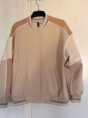 Beige bomberjacka från H&M - Snygg bomberjacka i olika beige nyanser från H&M. Jackan har vita kontrastsömmar, ribbade muddar och krage med randiga detaljer. Stängs med tryckknappar och har två fickor framtill. Perfekt för en avslappnad och trendig look.