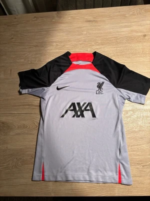 Liverpool tränings t-shirt Nike - Snygg Liverpool FC tränings t-shirt från Nike med Dri-FIT material. Tröjan är ljusgrå med svarta ärmar och röda detaljer vid hals och sidor. Tryck med AXA och LFC-logga på bröstet. Perfekt för fotbollsträning eller gymmet. Passar dog som är runt 165