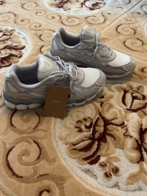 Gråvita ASICS GEL NYC sneakers - Snygga ASICS GEL sneakers i grått och vitt med mesh- och syntetpaneler. Skorna har en chunky sula och klassisk snörning. Perfekta för dig som gillar sportig och stilren look. ASICS-logga på sidan och GEL-detalj bak.