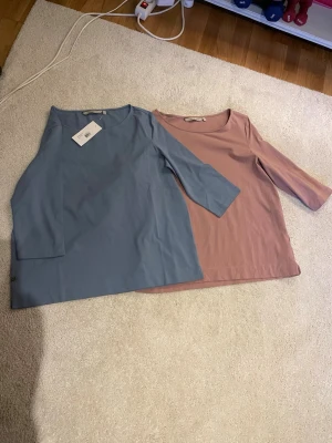 Blå och rosa toppar från House of Lola - Två stilrena toppar från House of Lola, en i ljusblått och en i dämpad rosa. Båda har rund halsringning och trekvartsärm. Perfekta att matcha med jeans eller kjol för en enkel men snygg look.