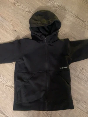 Svart ziphoodie från Sail Racing - Clean hoodie från Sail Racing med dragkedja framtill och två stora fickor. Huva med diskret logga och stilren design.