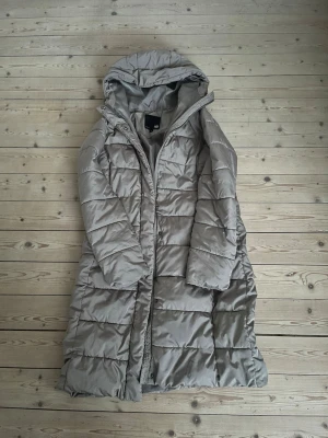 Beige lång dunjacka från 157 - Lång beige dunjacka från 157 med huva och dragkedja framtill. Jackan har quiltad design med breda kanaler och tryckknappar längs öppningen. Perfekt för kalla vinterdagar och riktigt skön att mysa in sig i.