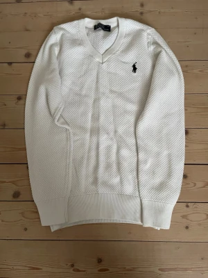 Vit stickad tröja från Polo Ralph Lauren - Vit stickad tröja med v-ringning från Polo Ralph Lauren. Tröjan har ett diskret strukturmönster och den klassiska svarta logotypen broderad på bröstet. Ribbstickade muddar vid ärmslut och nederkant. Perfekt för en stilren look. S