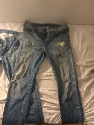 Blå slitna jeans från Replay - Säljer ett par blå jeans från Replay med slitningar och hål för en trendig, avslappnad look. Jeansen har knäppning fram och klassisk femficksmodell. Perfekta för dig som gillar en streetstyle vibe och vill ha ett par riktigt snygga jeans.