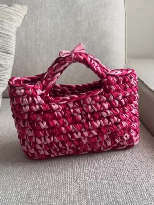 Handgjord rosa och röd virkad handväska - Unik handväska i virkad design med breda band i olika nyanser av rosa och rött. Väskan har en rektangulär form, dubbla handtag och en liten rosett på toppen. Perfekt för dig som vill sticka ut med en färgglad accessoar.