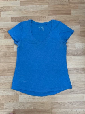 Blå v-ringad t-shirt från Lindex - En enkel och snygg blå t-shirt från Lindex med v-ringning och korta ärmar. Tillverkad i mjuk bomull som känns skön mot huden. Perfekt till jeans eller shorts för en avslappnad look.
