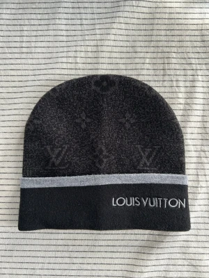 Svart mössa från Louis Vuitton - Snygg svart mössa från Louis Vuitton med diskret monogrammönster och grå rand längs kanten. Broderad logga framtill. Perfekt accessoar för dig som vill ha en exklusiv och stilren look. Mjuk och stretchig kvalitet.