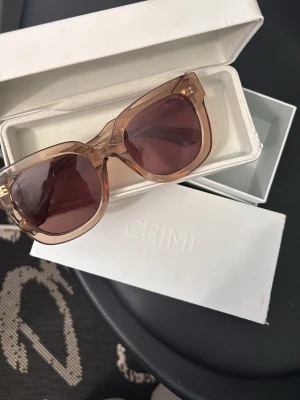 Bruna solglasögon från CHIMI - Snygga solglasögon från CHIMI med transparent, beige-brunt båge och mörkbruna glas. Rektangulär form som ger en modern vibe. Kommer med originaletui och putsduk. Perfekta för soliga dagar och trendiga outfits.
