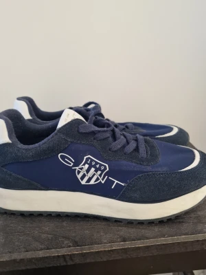 Marinblå sneakers från GANT - Snygga marinblå sneakers från GANT med vit logga och detaljer. Ovandelen är i mocka och textil, med vita inslag på sulan och hälen. Klassisk låg modell med snörning och bekväm sula. Perfekta för en sportig och stilren look.