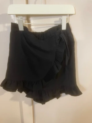 Svarta volangshorts från ONLY - Jag har storleken 164, och dom passar jätte bra på mig. Säljer ett par svarta shorts från ONLY med volangkant och omlottdesign. Shortsen har en mjuk resår i midjan och knyt i sidan för extra detalj. Perfekta för sommarens alla tillfällen och riktigt sköna att bära.