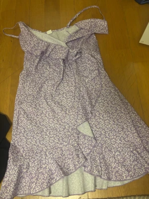 Ljuslila blommig omlottklänning - Söt ljuslila omlottklänning med vita små blommor, tunna axelband och volangdetaljer längs nederkanten. Klänningen har en feminin v-ringning och knyt i midjan för en snygg passform. Perfekt för varma dagar och somriga tillfällen.