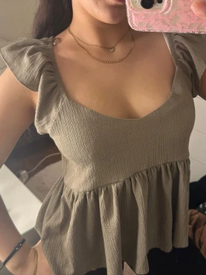 Beigebrun blus med volangärmar - Säljer en beige blus med volangärmar och v-ringning. Toppen har en luftig passform med rynkad nederdel och är tillverkad i ett lätt, ribbat material. Perfekt för dig som gillar en feminin och trendig stil.