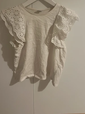 Vit topp med broderade volangärmar Lindex - Supersöt vit topp från Lindex med breda volangärmar i broderad spets. Toppen har rund halsringning och är tillverkad i mjuk bomull. Perfekt för dig som vill ha en söt och trendig look med extra detaljer på ärmarna.