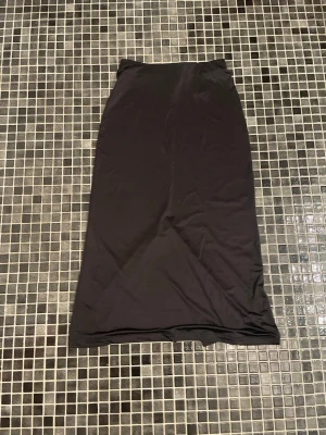 Svart långkjol med slits - En stilren svart långkjol i mjukt material med rak passform. Kjolen har en diskret slits baktill som ger extra rörelsefrihet. Perfekt att matcha med en crop top eller oversized tröja för en trendig look.