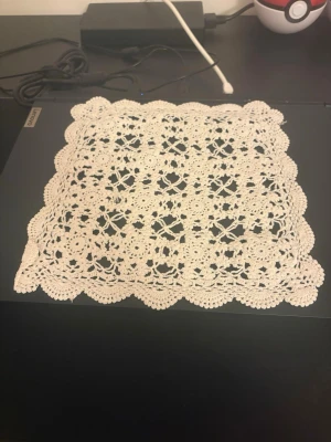 Virkad vit duk med spetsmönster - Säljer en handgjord virkad duk i vitt med detaljerat spetsmönster och vågiga kanter. Duken är fyrkantig och passar perfekt som dekoration på bord eller hylla. Materialet är tunt garn som ger ett klassiskt och elegant intryck.