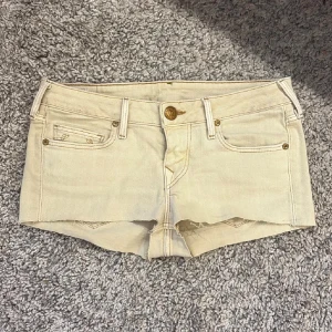 Beige denimshorts från True Religion - Säljer ett par ljusbeige denimshorts från True Religion med rå kant nertill, klassiska fem fickor och guldfärgade knappar. Shortsen har låg midja och är perfekta för sommaren. Snygg broderad detalj på bakfickorna och läderpatch med logga bak. Klippt t shorts