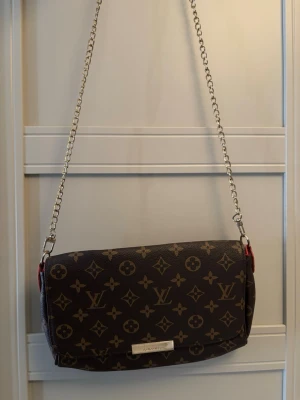  axelväska - Säljer en klassisk brun axelväska med ikoniskt monogrammönster i beige. Väskan har en kedjerem i silverfärg och detaljer i rött vid sidorna. Tillverkad i tåligt canvasmaterial med läderkänsla och har en metallbricka framtill med logotyp.