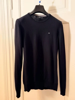 *BRA DEAL* stickad tröja från J.Lindeberg - Klassisk svart stickad tröja från J.Lindeberg med rund halsringning och diskret logga broderad på bröstet. Tröjan har lång ärm och ribbade muddar vid ärmslut och nederkant. Perfekt basplagg i garderoben.