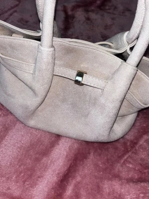 Beige mocka handväska med gulddetalj - Säljer en snygg beige handväska i mjuk mocka med guldfärgad metallknäppning. Väskan har dubbla handtag och en enkel, stilren design. Perfekt för dig som gillar neutrala färger och klassisk look. Tillhörande långt band medföljer. Aldrig använd 