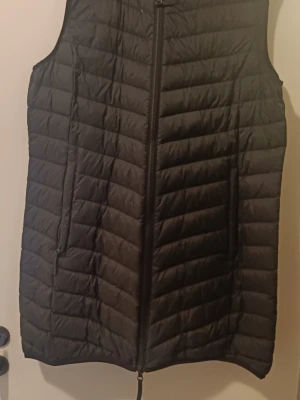 Svart lång väst med dragkedja - Snygg svart dunväst med och hel dragkedja framtill. Västen har två fickor på sidorna och en stilren, enkel look som passar till många outfits. Perfekt lager på lager-plagg för kyligare dagar.