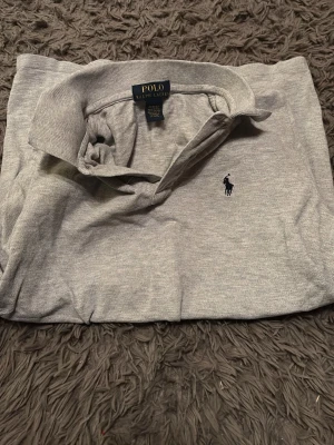 Grå pikétröja från Polo Ralph Lauren - Klassisk grå pikétröja från Polo Ralph Lauren med den ikoniska broderade loggan på bröstet. Tillverkad i mjuk bomull och har krage samt knappar vid halsen. Perfekt för en clean och stilren look.