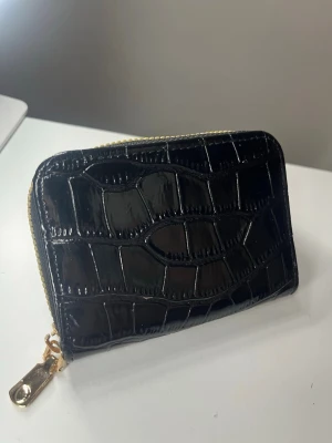 Svart plånbok från zara - Snygg svart plånbok i glansigt konstläder med krokopräglat mönster och guldfärgad dragkedja. Plånboken har flera fack på insidan för kort och sedlar. Perfekt för dig som vill ha en stilren och trendig accessoar.