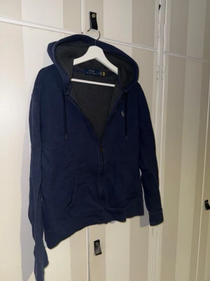 Marinblå hoodie från Polo Ralph Lauren - Snygg marinblå hoodie från Polo Ralph Lauren med dragkedja, huva med dragsko och två stora fickor framtill. Insidan är mjukt fodrad i grått och den klassiska loggan är broderad på bröstet. Perfekt för en chill och avslappnad stil.                                              Hör av er vid frågor!