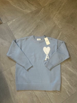 Ljusblå stickad tröja från Ami Paris - Ljusblå stickad tröja från Ami Paris med rund halsringning och ribbade muddar. På bröstet finns ett vitt hjärta med bokstaven 'A' under, vilket är Ami Paris ikoniska logga. Tröjan har en avslappnad passform och är perfekt för lager på lager.