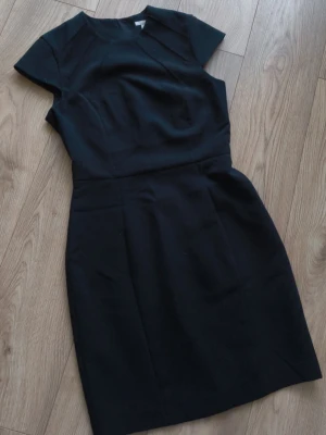 Svart kort klänning - En stilren svart klänning med korta ärmar och rund hals. Klänningen har en figurnära passform och dragkedja i ryggen. Perfekt för dig som gillar en klassisk look med enkel design.