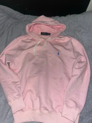 Rosa hoodie från Polo Ralph Lauren - Snygg ljusrosa hoodie från Polo Ralph Lauren med klassisk blå logga broderad på bröstet. Hoodien har huva med dragsko, magficka och ribbade muddar. Perfekt för en chill och avslappnad stil. Materialet är mjukt och skönt, troligen bomull.