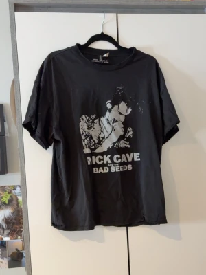 Svart band-tshirt Rick Cave Divided - Cool svart t-shirt från H&M Divided med tryck av Rick Cave and the Bad Seeds på framsidan. Oversized passform med korta ärmar och rund halsringning. Perfekt för dig som gillar bandmerch och vill ha en avslappnad stil.