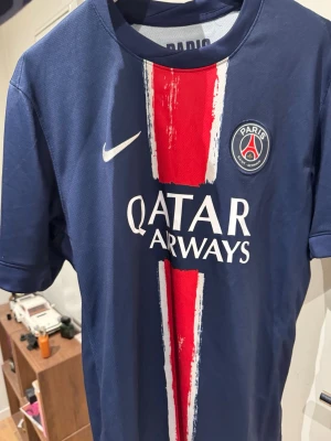 PSG matchtröja blå Nike - Snygg blå PSG matchtröja från Nike med röd och vit rand i mitten, Qatar Airways-tryck och klubbmärke på bröstet. Tillverkad i lätt och ventilerande material, perfekt för fotboll eller träning. Kortärmad och med klassisk rund hals.