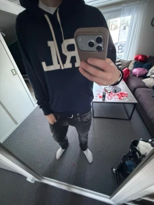 Mörkblå RL hoodie med huva - Snygg mörkblå hoodie från Ralph Lauren med stor vit RL-logga på bröstet. Hoodien har huva med vita snören och en klassisk känguruficka framtill. Perfekt för en chill och avslappnad stil. Materialet känns mjukt och bekvämt.