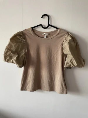 Beige topp med puffärmar från H&M - Trendig beige topp från H&M med stora puffärmar. Toppen har rund halsringning ärmar i ett krispigare material utvändigt men mjukt T-shirtmaterial inuti. 