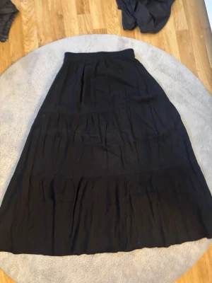 Svart långkjol i bomull - Säljer en svart långkjol med bred resår i midjan och lätt utställd passform. Kjolen har flera diskreta sömmar som ger en snygg struktur och faller mjukt. Perfekt att styla med sneakers eller boots för en chill look. Jätte skön men inte min typ av kjol! Använd få tal gånger!