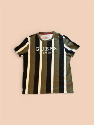 Randig t-shirt från Guess - Snygg randig t-shirt från Guess med breda vertikala ränder i svart, olivgrön och vit. Tryck med 'GUESS EST. 1981' på bröstet. Regular fit och klassisk rund hals. Perfekt för dig som gillar streetwear vibes.