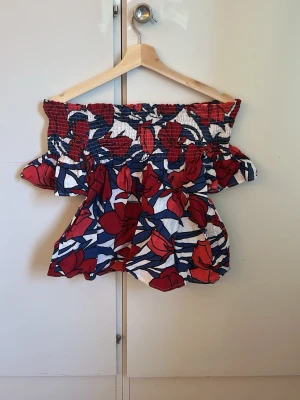 Offshoulder topp med blommigt mönster - Säljer en färgglad offshoulder topp med blommigt mönster i rött, blått och vitt. Toppen har smock upptill och volangdetaljer som ger en luftig och somrig känsla. Perfekt för dig som vill sticka ut med en unik och trendig stil. Köpte den utomlands så vet inte vilken märke den är ifrån.
