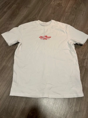 Vit t-shirt med röd brodyr - Snygg vit t-shirt med röd broderad text på bröstet där det står 'Los Angeles Beverly Hills Calif. MCMXCVII'. Klassisk rund hals och korta ärmar. Perfekt för en avslappnad och trendig look.