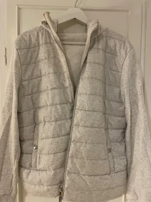 Ljusgrå pufferjacka med dragkedja - Säljer en ljusgrå pufferjacka med quiltad front och släta ärmar. Jackan har hel dragkedja, två fickor med tryckknappar och hög krage. Perfekt för kalla vinterdagar och har en modern, enkel design.