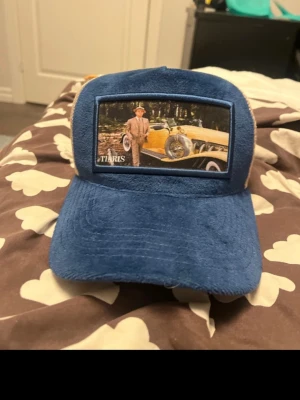 Blå vintageinspirerad keps från Tiaris - Säljer en blå keps från Tiaris med beige mesh baktill och en unik patch framtill med vintagebil och man i kostym. Kepsen har böjd skärm och mjuk plyschliknande yta. Justerbar baktill för perfekt passform. Perfekt för dig som gillar retrostil.