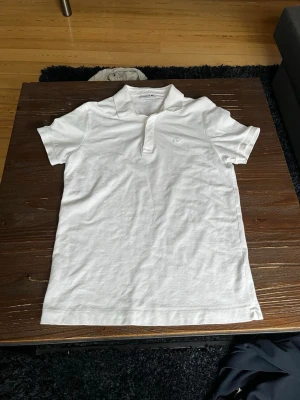 Vit pikétröja från Lacoste - Klassisk vit pikétröja från Lacoste med krage och knappslå framtill. Tröjan har korta ärmar och den ikoniska krokodilloggan broderad på bröstet. Regular fit och tillverkad i bomull för en bekväm känsla. ALDRIG ANVÄND!!