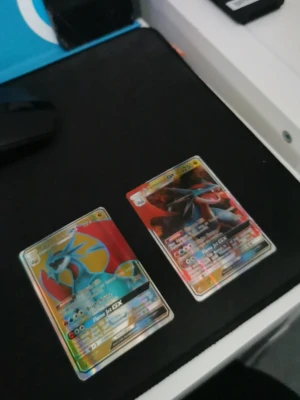 Två Pokémon GX samlarkort - Säljer två Pokémon GX samlarkort med färgstarka motiv och holografisk finish. Ena kortet har blå och orange färger med en drakliknande Pokémon, det andra har röd och svart design med en annan Pokémon. Perfekta för samlare eller Pokémonfans.