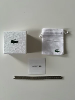 Silverfärgat armband från Lacoste - Elegant armband från Lacoste i rostfritt stål med stilren och tidlös design.   Den borstade metallen tillsammans med den diskreta krokodilloggan ger ett sportigt men sofistikerat uttryck som passar både vardag och mer uppklädda tillfällen.    Levereras med ask, påse och kort enligt bilderna, vilket gör det extra fint även som present.    En stilfull accessoar som lyfter en enkel outfit och fungerar året runt.   Skickas snabbt och packas noggrant.