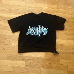 CROPPED Svart AMIRI t-shirt  - Svart t-shirt med stort grafiskt AMIRI-tryck i vitt och blått framtill. Modellen är kortärmad med rund hals och har en loose passform. Perfekt för en avslappnad och trendig stil.